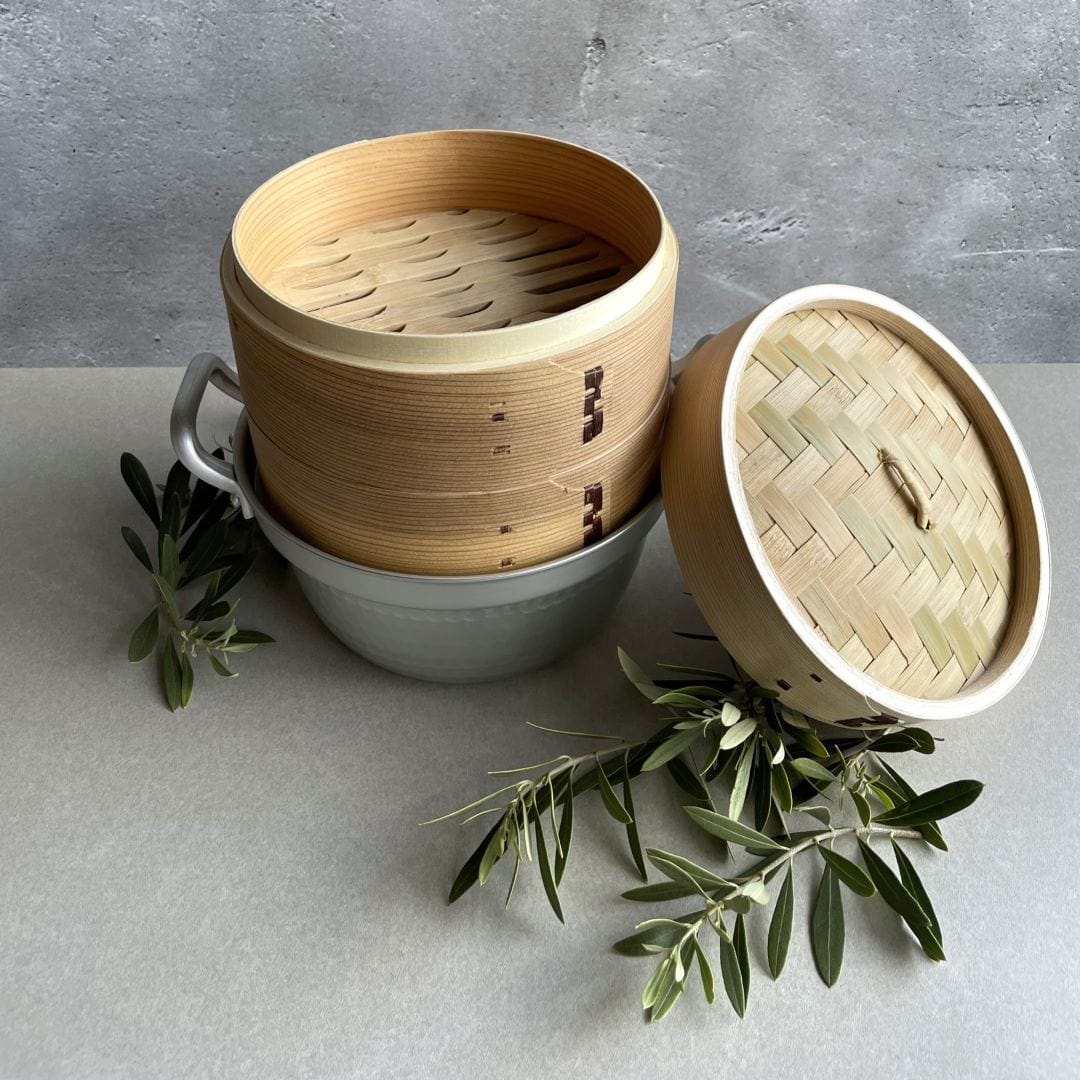 A minimalist setup with a grey wall in the background and a grey tabletop in the foreground. A brown bamboo steamer basket with its lid open is placed in the middle of the table, revealing its contents. Two olive branches are symmetrically arranged around the basket, adding a touch of greenery to the neutral color scheme. The composition evokes a sense of simplicity and natural beauty.
