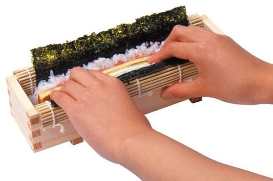 Two hands on a sushi roll maker with seaweed leaf inside a sushi maker box with rice and fish inside