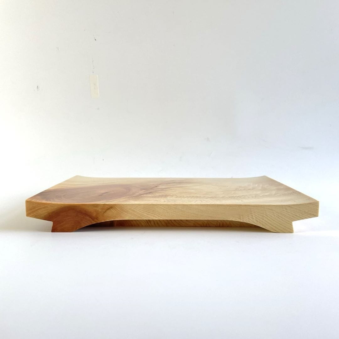 An image of a wooden sushi tray captured in a horizontal view. The tray is placed on a white surface, and the surrounding environment is also white, creating a clean and minimalistic look. The wooden texture of the tray is prominent, featuring natural grains and patterns. The sushi pieces are not visible in this image, but the tray&