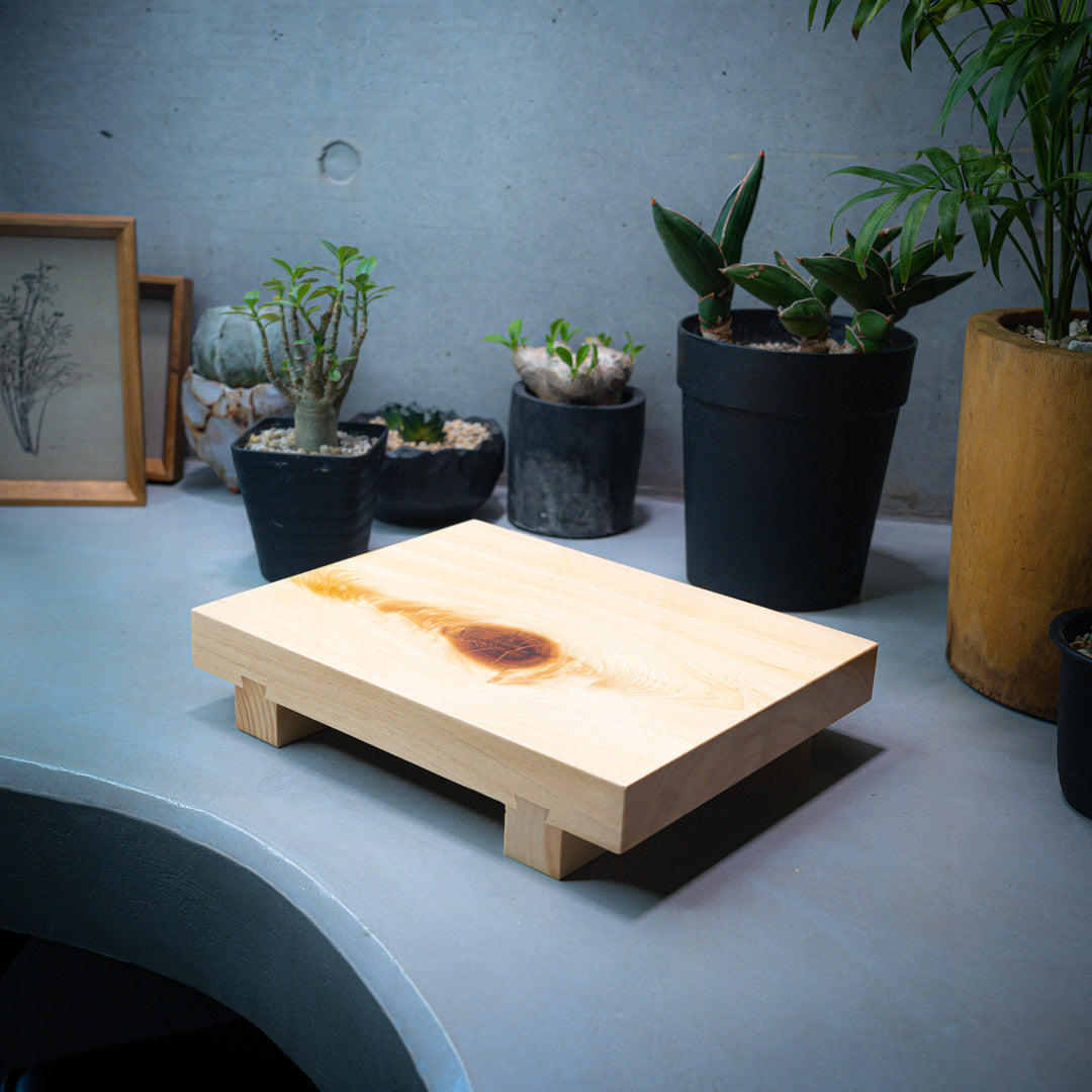 An overhead view of a wooden sushi tray placed on a gray kitchen counter. In the background, four small plants are visible, adding a touch of greenery to the scene. The backdrop consists of a plain gray wall, providing a simple and neutral background to the composition.