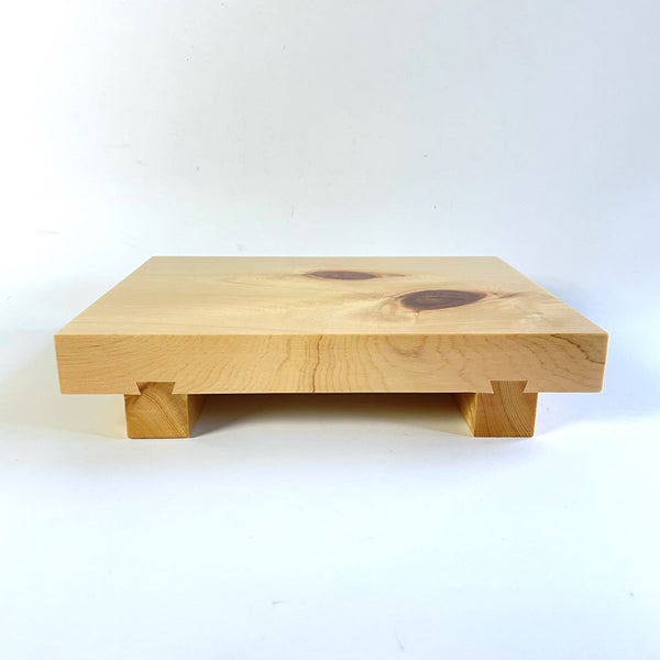 An image of a wooden sushi tray captured in a horizontal view. The tray is placed on a white surface, and the surrounding environment is also white, creating a clean and minimalistic look. The wooden texture of the tray is prominent, featuring natural grains and patterns. The sushi pieces are not visible in this image, but the tray's simple and elegant design is the focal point of the composition.