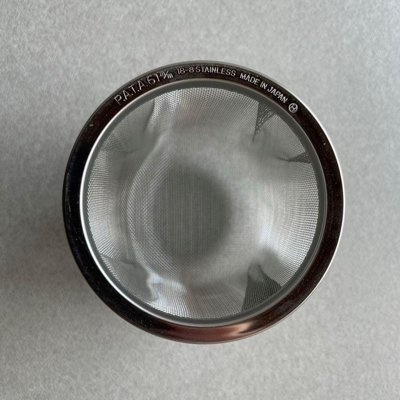 This is an upper view of a stainless steel infuser placed in the middle of a grey table. The infuser has a mesh-like structure with evenly spaced holes and a circular shape.