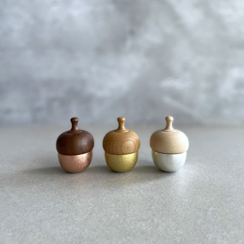 Three handcrafted Healing Bells standing in a row on a grey surface. The first one has a pink color, the second one is goldish, and the last one is white-grey. Each bell has an acorn shape with a wooden top and a brass pendulum tucked inside. The bells are displayed in the middle of a grey room.