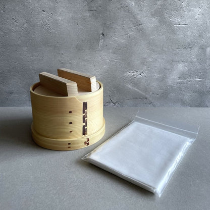 A closed lid wooden container, made of uncoated sawara cypress wood, with a white sheet next to it, used for making tofu. The container sits in the middle of a grey room.