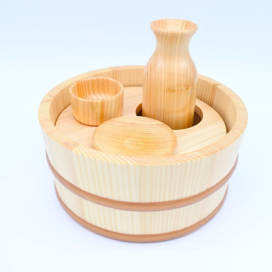 A wooden set of sake drinking devices, including a tub, a bottle, a cup, and a plate, placed in a white room.
