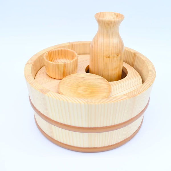 A wooden set of sake drinking devices, including a tub, a bottle, a cup, and a plate, placed in a white room.